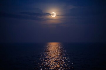 A full moon casts a shimmering reflection on the dark ocean, with a few clouds partially obscuring the night sky.
