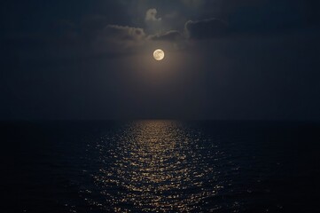 A full moon casts a shimmering reflection on the dark ocean, with a few clouds partially obscuring the night sky.