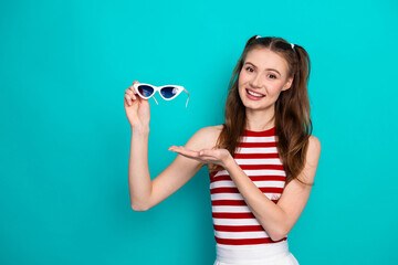 Young cheerful woman displaying stylish sunglasses with a vibrant smile on a trendy turquoise...