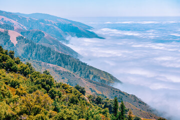 Majestic Big Sur mountains in California USA covered with lush greenery, rolling ridges, and dramatic marine layer clouds, creating a breathtaking coastal landscape above the Pacific Ocean