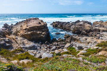 Rocky shoreline along 17 Mile Drive near Monterey, California. Waves crash against rugged stones, with coastal plants adding color under a bright sky on this famous scenic route by the Pacific Ocean