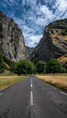 A long, paved road winds through a valley, bordered by towering, rugged mountains under a dramatic sky filled with fluffy clouds.