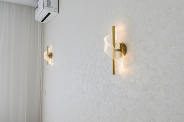 A minimalist interior with two golden sconces on a textured white wall, near a white curtain and air conditioning unit