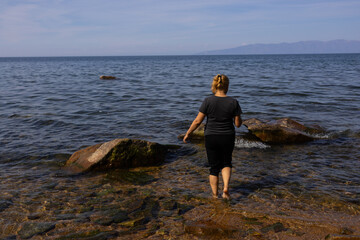 A white woman with blonde hair stands on a rocky shore, looking at calm water and distant mountains.