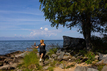 Obraz premium A white woman with blonde hair stands on the rocky shore of Lake Baikal. She holds a phone in her hands and looks at the water. Mountains are visible in the background.