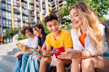 Happy friends in casual fashion sharing fun moments on devices outdoors in city during sunny day together