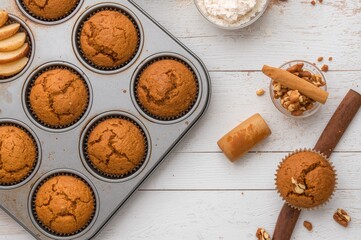 Fototapeta premium Cinnamon-spiced apple muffins topped with chopped walnuts and apple pieces in a metal baking tray on a white wooden surface. Freshly baked homemade dessert with baking essentials. Overhead shot.