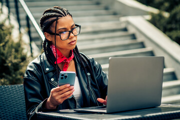 Young woman working outdoors with laptop and phone while enjoying leisure time in a casual urban...
