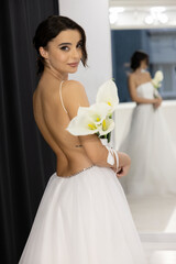 Stylish bride in elegant white wedding dress posing with lilies indoors