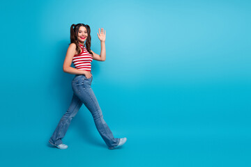 Stylish young woman waving with a smile against vibrant background wearing casual denim and striped top