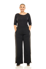 Studio shot of bald smiling woman in black top and wide-leg pants standing full length on white background.