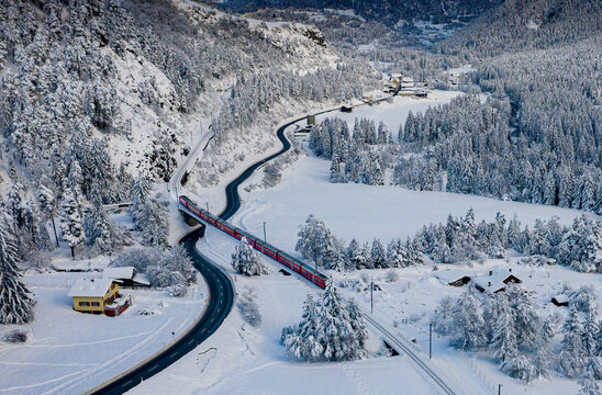 Aerial view of a vibrant red train gracefully winding through a snow-laden valley, juxtaposed against the stark white landscape, Filisur, Switzerland.