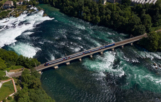 Aerial view of a train crossing a bridge over the churning, turquoise Rhine River, with the powerful Rhine Falls cascading nearby, Filisur, Switzerland.