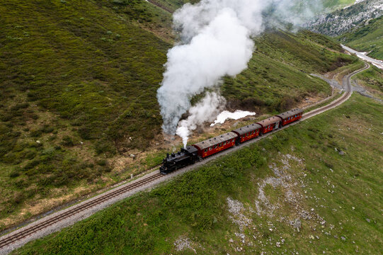 Aerial view of a vintage steam train winding through the verdant, steep slopes, plumes of white smoke contrasting against the green landscape, Furka Pass, Switzerland.