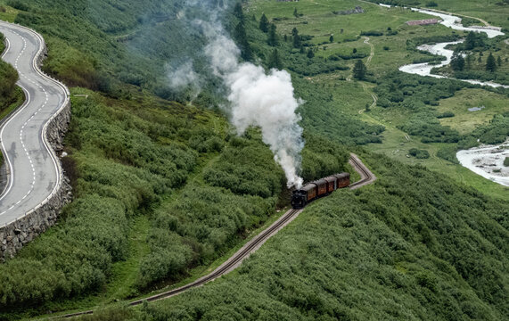 Aerial view of a vintage train puffs white clouds while winding through lush green hills alongside a snaking river and a winding road, Furka Pass, Switzerland.