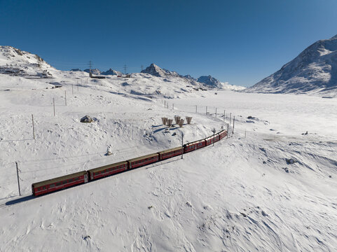 Aerial view of a deep red train winding through the stark white, snow-covered mountains under a crisp blue sky, Bernina Express Switzerland.