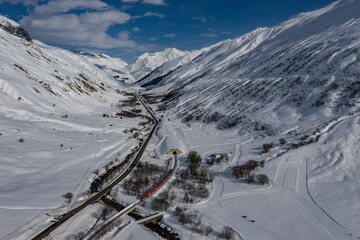 Aerial view of a red train snaking through the snow-covered valley, emerging from a tunnel beneath the towering, snow-capped Alps, Filisur, Switzerland.