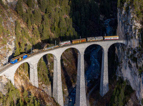 Aerial view of a train traversing the majestic Landwasser Viaduct, an architectural marvel against a backdrop of rugged cliffs and a winding river, Filisur, Switzerland.