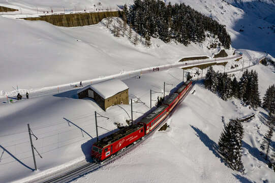 Aerial view of a red train slicing through a brilliant white snowscape near an old stone structure, contrasting against the dark green pines, Filisur, Switzerland.