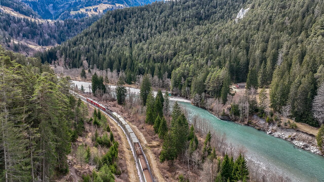 Aerial view of a red train winding through the dense forest alongside a turquoise river, a stunning contrast of colors and textures, Rhine River, Switzerland.