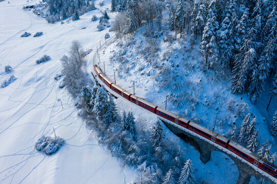Aerial view of a vibrant red train slicing through the snow-laden landscape near the frosty, snow-capped trees, Filisur, Switzerland.
