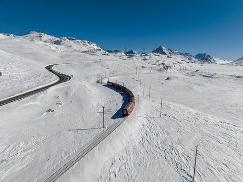 Aerial view of a red train curving through a snow-laden landscape, contrasting against the stark white expanse under a clear blue sky, Bernina Express Switzerland.
