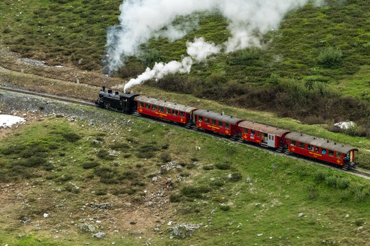 Aerial view of a vintage red train weaving through green hills as smoke billows, creating a striking contrast against the landscape, Furka Pass, Switzerland.