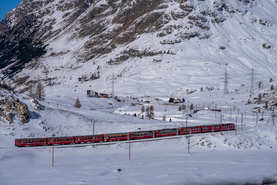 Aerial view of a vibrant red train slicing through the pristine white snow-covered landscape near the majestic mountains, Filisur, Switzerland.