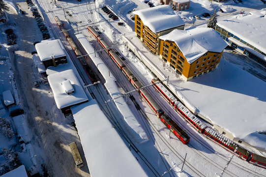 Aerial view of red trains stand out vividly against the pristine white snow covering tracks and buildings, creating a striking winter landscape, Andermatt, Switzerland.