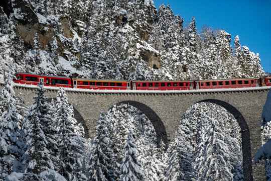 Aerial view of a vibrant red train traverses a snow-laden stone arch bridge amidst the frosted evergreens and rocky cliffs, Filisur, Switzerland.