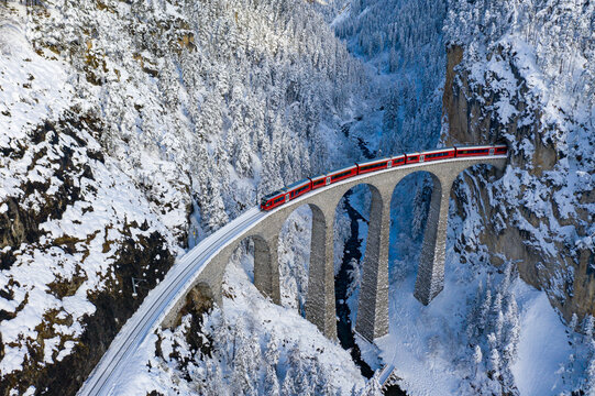 Aerial view of a red train gracefully traversing the snow-laden Landwasser Viaduct, a stark contrast against the frosted landscape, Filisur, Switzerland.