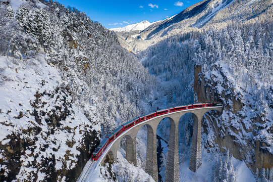 Aerial view of the majestic Landwasser Viaduct curving through a snow-laden valley, the red train contrasting against the stark white landscape, Filisur, Switzerland.