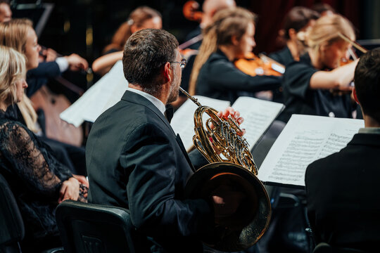 A focused French horn player leads an orchestra in a dramatic, well-lit concert. Musicians in formal wear perform with intense concentration, surrounded by sheet music and rich acoustic ambiance