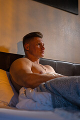 man sits upright in bed with his bare chest exposed gazing thoughtfully into the distance. The evening light casts a warm glow creating a tranquil atmosphere in the room.