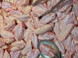 A pile of raw, pale pink chicken middle wings with skin, glistening and moist, gathered in a display container with a metal spoon and tongs resting on top.