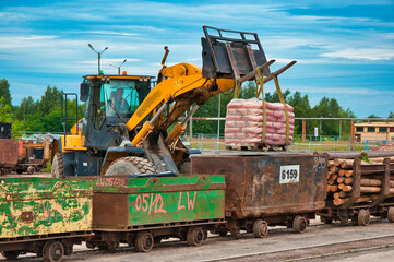  Jasnożółta koparka hydrauliczna ładuje paletę z materiałem w workach na zardzewiały wagon kolejowy na terenie czynnego składu drewna. W tle widać kłody drewna, szyny i inny sprzęt budowlany. © Pol Badger