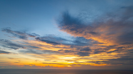 A breathtaking sunset paints the sky in vivid hues of orange and blue over a coastal town, with lush green hills framing the scene. A distant hilltop structure adds a serene tranquil landscape.