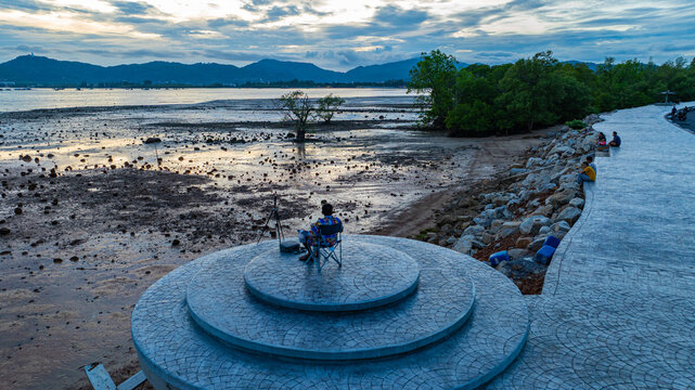 A man sits alone eating in a folding chair on a stone platform, overlooking the rocky intertidal flats, camera at his side, and the peaceful sea in the distance. A quiet moment of solitude at sunset.