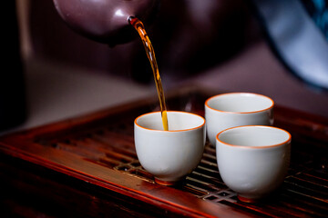 Traditional tea pouring scene with ceramic teacups on a wooden tray. Warm lighting, cultural...