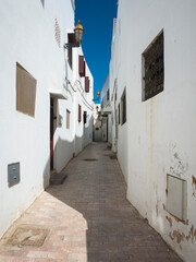 Quiet narrow alley in Rabat Kasbah in Morocco, with whitewashed walls and stone pavement. Small shuttered windows under blue sky. Half in bright sunlight, half in shadow - completely empty