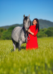 Woman with dark hair and bright red dress walking her Arabian gray horse in green field on sunny day, smiling as animal eats grass