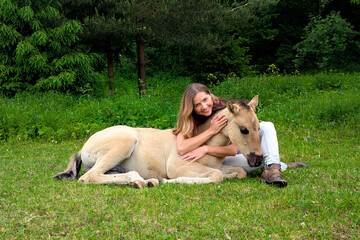 Young woman sitting on ground next to dunn color small horse foal, another animal near in background