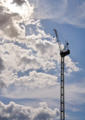 Looking up tall construction crane, sun shines through clouds in background