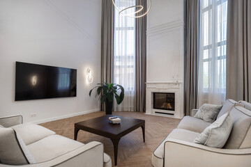 Spacious living room features stylish furniture, a large screen TV, and a cozy fireplace. Sunlight streams through tall windows, creating a warm atmosphere