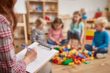 In a bright classroom, a teacher takes notes while four children play with colorful building blocks on the floor, promoting creativity and teamwork among them