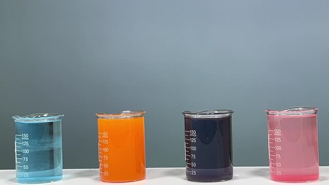 Four laboratory glass beakers with colorful liquid solutions in blue, orange, black, and pink on white surface with plain gray background
