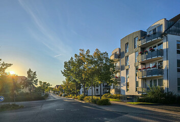 quiet residential street with modern apartment buildings on the right side, bathed in the warm light of the setting sun. Trees line the street, casting long shadows, while balconies with flowers