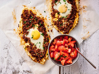 Two delicious pide topped with minced meat and egg are presented on parchment paper, accompanied by a bowl of fresh tomato salad. This meal highlights authentic Turkish flavors