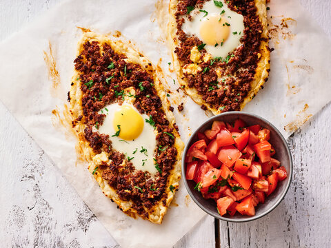 Pide, a traditional Turkish flatbread pizza, is topped with spiced meat and includes an egg. It is served alongside a fresh tomato salad, showcasing vibrant colors and flavors
