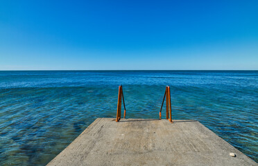Pier with rusty railings for descent into the sea, clear calm weather with a view of the calm sea, background idea about marine ecosystems and human impact